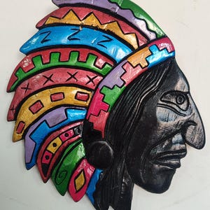 以下が含まれることがあります： 木製の壁掛けで、横顔のネイティブアメリカンのチーフを描いています。彫刻は黒い顔と、赤、青、緑、黄、紫のアクセントが付いたカラフルな頭飾りを特徴としています。アートワークは白い表面に取り付けられています。
