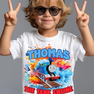 Puede incluir: Camiseta blanca con el nombre "THOMAS" en letras azules de burbujas y una colorida ilustración de Thomas the Tank Engine. El texto "THE TANK ENGINE" está debajo de la ilustración del tren en rojo.