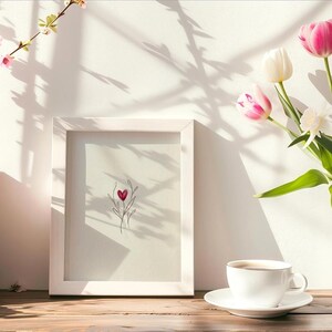 May include: A still life arrangement featuring a framed artwork with a heart design, a cup of coffee, books, and a vase of tulips. The scene is bathed in sunlight, creating shadows on the white wall and wooden surface.