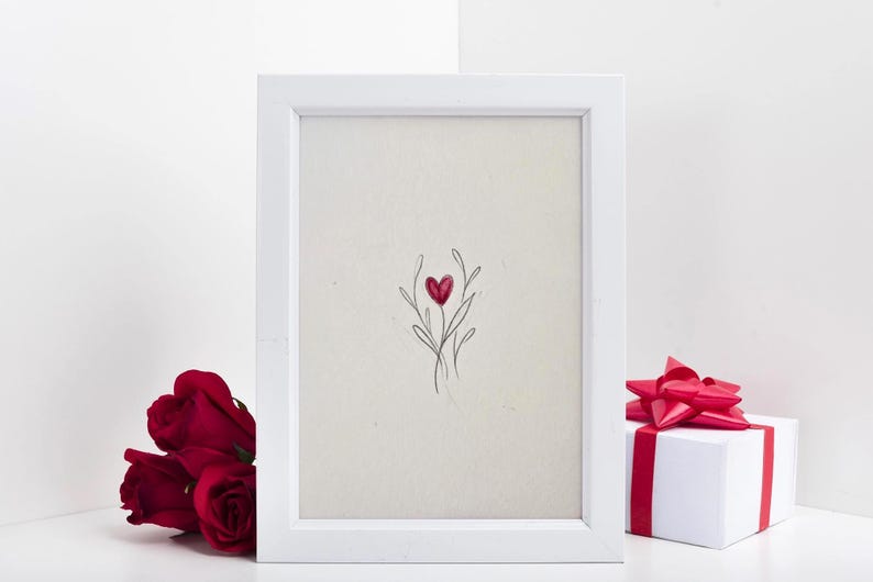 May include: A white framed art print featuring a red heart-shaped floral design. Beside the frame are three red roses and a white gift box with a red ribbon. The artwork is on a white background.