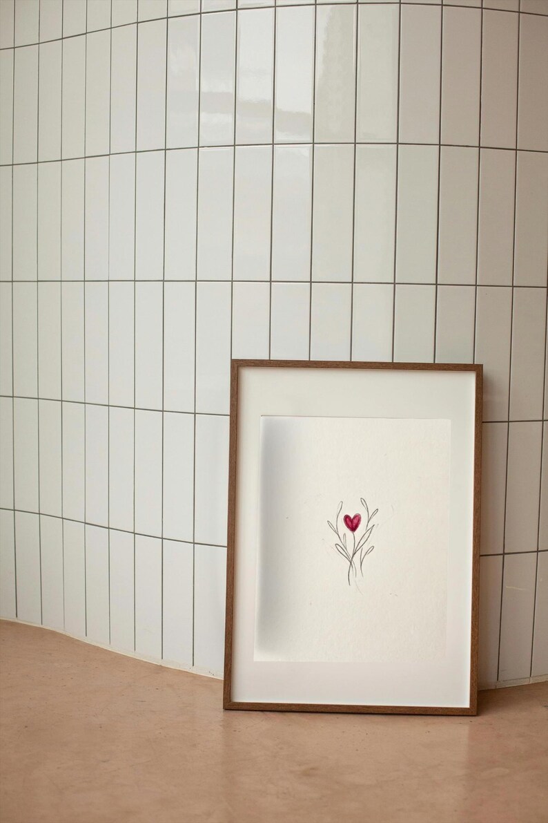 May include: A framed artwork featuring a minimalist illustration of a red heart with delicate lines. The artwork is set against a white background within a wooden frame. The background is a white tiled wall.