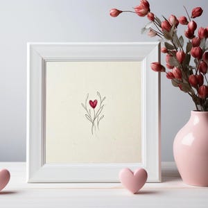May include: A white framed artwork with a minimalist heart design in red and black. A pink vase with red flowers and two pink heart-shaped decorations are on a white surface, creating a romantic theme.