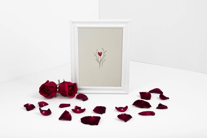 May include: A white framed art print with a minimalist design of a red heart and floral elements. Two red roses and scattered petals surround the frame on a white surface. The image has a romantic theme.