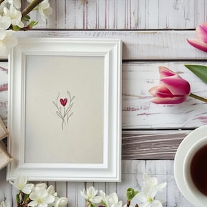 May include: A white framed art print featuring a red heart design, alongside a cup of tea, tulips, and books. The image is set on a white wooden surface, with floral accents.