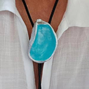 May include: A turquoise and silver bolo tie. The pendant is a shell shape with a bright turquoise center and a silver border. The black cord has decorative silver tips. The tie is worn over a white button-down shirt.