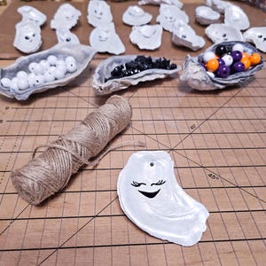 May include: A collection of painted oyster shells with smiling faces, various beads, and a spool of twine on a wooden surface. The shells are white with black facial details. Some shells are filled with white, black, purple, and orange beads.