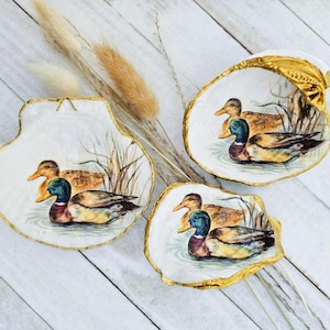 May include: Three decorative seashell dishes with hand-painted duck scenes. Each shell features a white interior with a gold-painted rim. The duck illustrations include a male mallard with a green head and a brown hen.