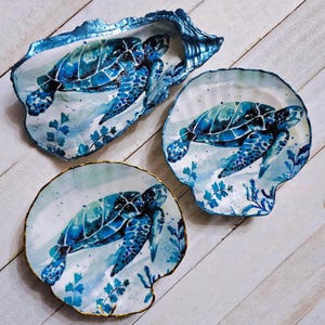 May include: Three decorative seashell dishes with hand-painted sea turtles. The turtles are depicted in shades of blue and teal, with intricate details. The shells have a white interior and are accented with blue and gold trim, creating a coastal theme.