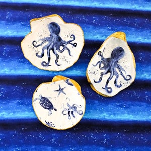May include: Three white seashell-shaped dishes with blue and gold accents. The dishes are decorated with painted images of an octopus, a turtle, and a starfish.