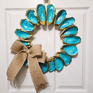 May include: A decorative wreath made of painted oyster shells. The shells are painted a vibrant turquoise blue with gold edges, arranged in a circular shape. A large burlap bow is attached at the bottom. The wreath hangs on a white door.