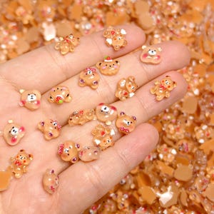 May include: A collection of small, translucent brown bear-shaped charms with various decorative elements like bows and strawberries. The charms are held in a hand, with a background of similar charms.