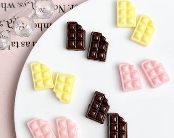 May include: A collection of miniature chocolate bar charms in various colors, including dark brown, yellow, and pink. The charms are arranged on a white surface, with a few pieces appearing to have a bite taken out of them.