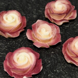 May include: Five sugar paste roses in shades of pink and white. The roses are arranged on a black textured surface.