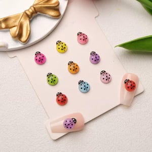 May include: A collection of colorful ladybug nail art decorations. The ladybugs are in various colors, including red, pink, yellow, green, orange, purple, and blue, each with black spots. Two nails are decorated with ladybugs. A gold bow is in the background.