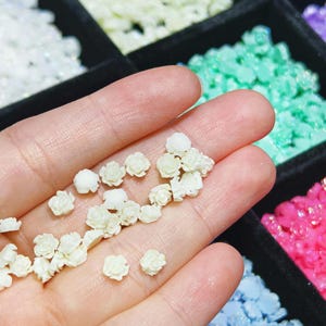 May include: A hand holding a collection of small, white, pearl-like flower embellishments. The flowers are in various sizes and shapes, with a slightly iridescent finish.