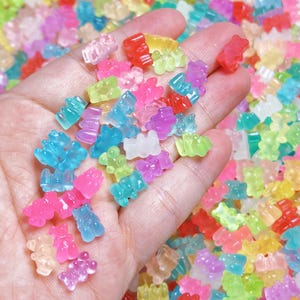 May include: A collection of small, translucent gummy bear-shaped charms in a variety of vibrant colors including red, blue, green, pink, purple, and yellow. The charms have a smooth, glossy finish and are scattered in a pile and held in a palm.