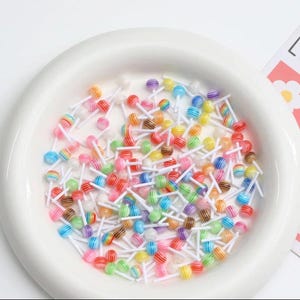 May include: A collection of miniature, colorful lollipops with striped designs on white sticks. The lollipops are in a white, shallow dish. The colors include red, orange, yellow, green, blue, purple, and brown.