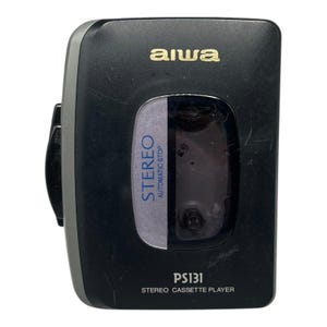 Könnte beinhalten: Ein schwarzer Aiwa PS131 Stereo-Kassettenrekorder mit grauer Seitenblende. Die Vorderseite verfügt über ein zentrales Sichtfenster für die Kassette, beschriftet mit dem blauen Text STEREO AUTOMATIC STOP sowie dem Modellnamen PS131 STEREO CASSETTE PLAYER.