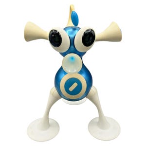 Könnte beinhalten: Eine blau-weiße Kunststoff-Spielfigur mit rundem Körper, zwei großen schwarzen Augen und zwei hornartigen Vorsprüngen an den Seiten. Sie verfügt über ein zentrales kreisförmiges Emblem, eine kleine Antenne oben und zwei weiße Beine mit Saugfüßen.