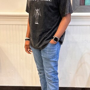 May include: A person wearing a black t-shirt with the text "SANTA NOIRE" and a white angel graphic. They are also wearing blue jeans and light brown sneakers.