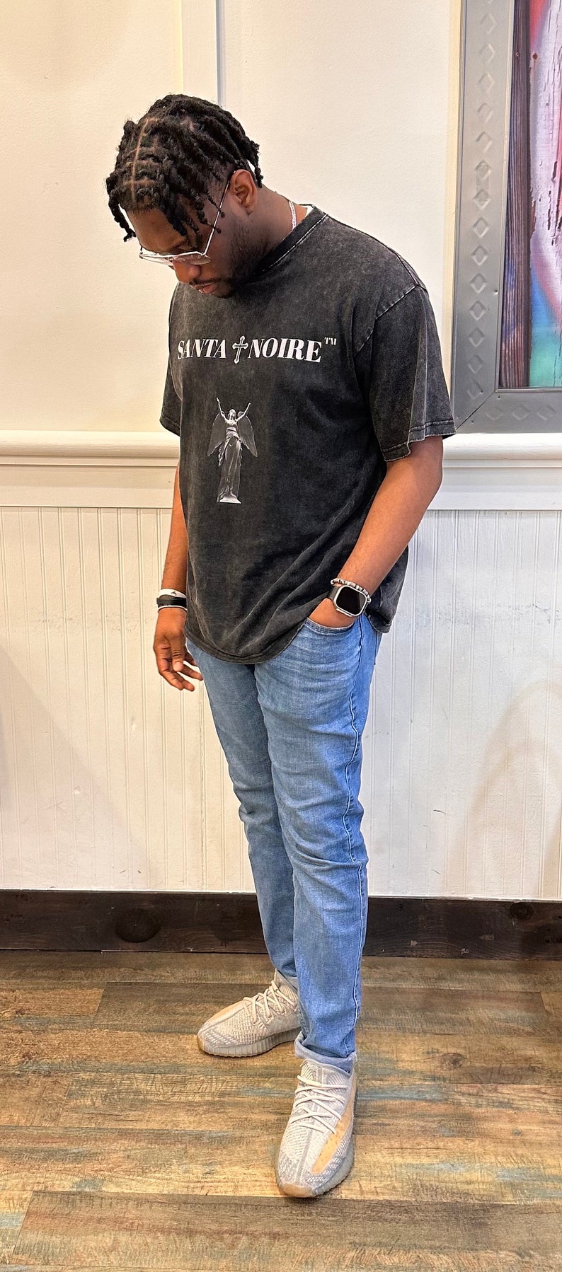 May include: A person wearing a black t-shirt with the text "SANTA NOIRE" and a white angel graphic. They are also wearing blue jeans and light brown sneakers.