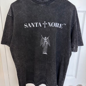 May include: Black t-shirt with a distressed look featuring a white graphic of an angel with wings and a cross above the text "SANTA NOIRE TM"