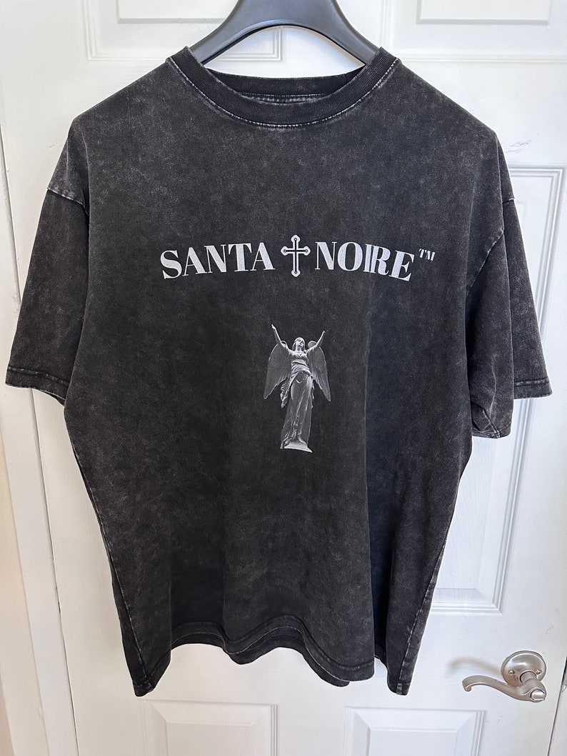May include: Black t-shirt with a distressed look featuring a white graphic of an angel with wings and a cross above the text "SANTA NOIRE TM"