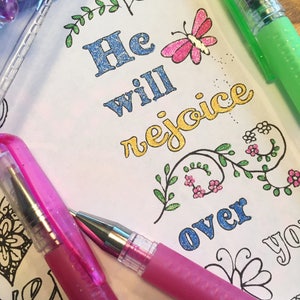 Puede incluir: Primer plano de una p&aacute;gina de libro para colorear con el texto "He will rejoice over you". La p&aacute;gina est&aacute; decorada con flores, mariposas y purpurina. Dos bol&iacute;grafos de gel rosa est&aacute;n sobre la p&aacute;gina.