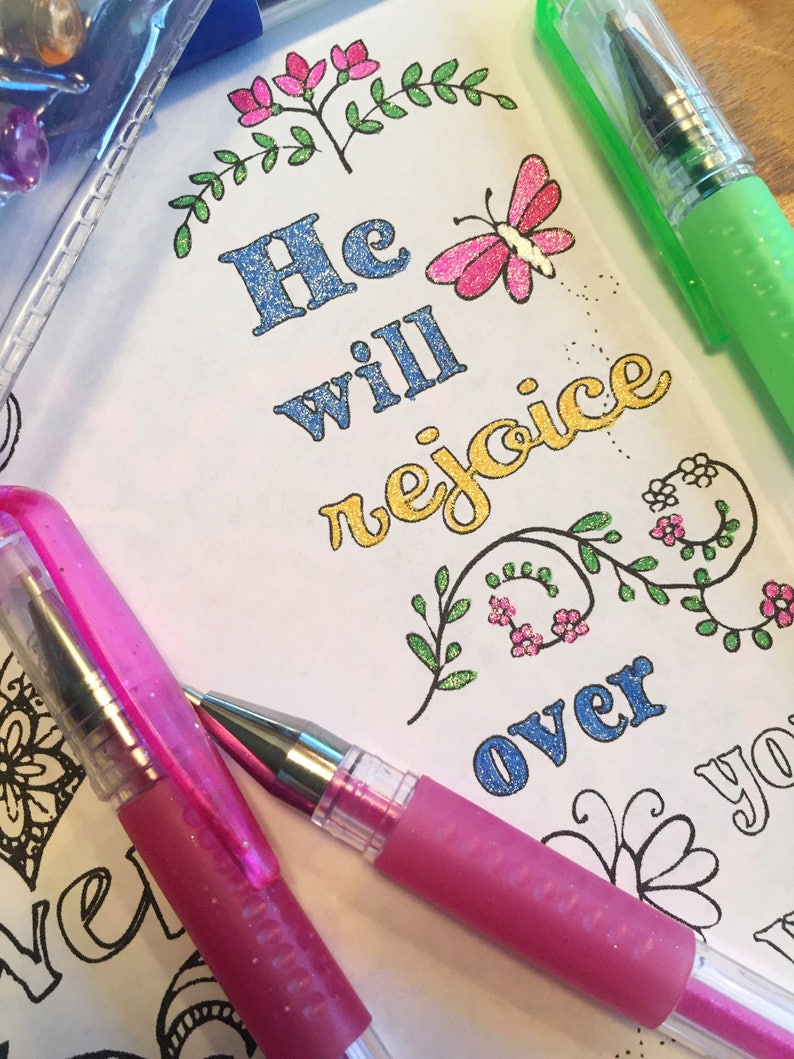 Puede incluir: Primer plano de una p&aacute;gina de libro para colorear con el texto "He will rejoice over you". La p&aacute;gina est&aacute; decorada con flores, mariposas y purpurina. Dos bol&iacute;grafos de gel rosa est&aacute;n sobre la p&aacute;gina.