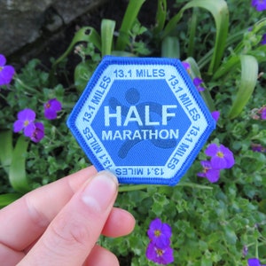 May include: A blue and light blue hexagonal patch with the words "HALF MARATHON" and "13.1 MILES". The patch features a runner graphic. The patch is held in front of a background of green foliage and purple flowers.