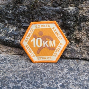 May include: Hexagonal orange and cream patch with the text "6.2 MILES" around the border. The centre features a running figure and the text "10KM". The patch is likely for a 10-kilometre race or running event.
