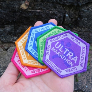 May include: A selection of colourful, hexagonal patches with embroidered text indicating running distances. Patches include distances like 3.1, 6.2, 13.1, and 26.2 miles, and the words "Ultra Marathon". The patches are in shades of pink, orange, blue, green, and purple.