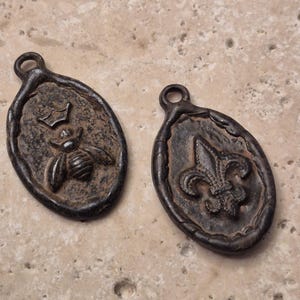 May include: Two oval-shaped metal pendants with a dark, weathered finish. One pendant features a raised bee icon topped with a small crown, while the other displays a raised fleur-de-lis emblem. Both pieces have a small loop at the top for hanging.