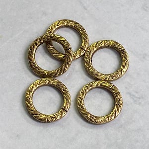 May include: Five gold-colored metal rings with a textured, rope-like design. The rings are circular and have a slightly aged appearance. The background is a light, marbled surface.