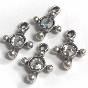 May include: Four silver-toned cross charms with a central clear crystal stone and four smaller ball accents. Each charm has a loop at the top for attaching to jewelry or other crafts. The charms are on a white background.
