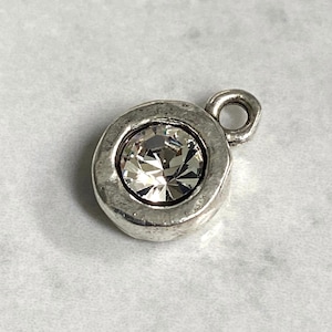 May include: A silver-toned charm with a large, clear crystal center. The charm has a textured, circular frame and a small loop for attachment. The crystal sparkles, reflecting light. The charm is likely for jewelry making or embellishment.