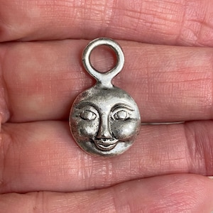 May include: A silver-toned charm in the shape of a smiling moon face. The charm has a loop at the top for attaching to a necklace or bracelet. The moon has detailed facial features, including eyes, a nose, and a smiling mouth.