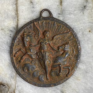 May include: A round, antique-style metal pendant with a loop at the top. The pendant features a central winged figure flanked by two horse-like creatures. The design is in relief, with a weathered, bronze-like finish.