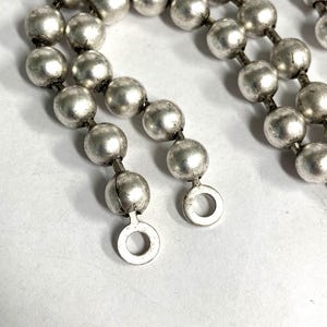 May include: Close-up of a silver-toned necklace featuring multiple strands of round, metallic beads. The beads are connected by small links, and the necklace ends with circular clasps. The jewelry has a vintage or antique appearance.