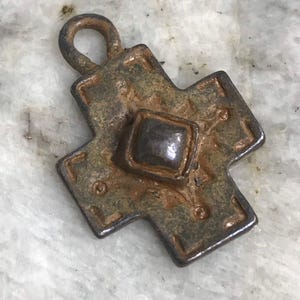 May include: A weathered, bronze-colored cross pendant with a square center stone. The cross has a loop at the top for hanging and features decorative detailing. The metal has a rustic, aged appearance.