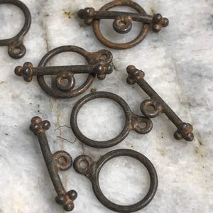 May include: Close-up of several antique bronze toggle clasps. The clasps feature a ring and a bar with decorative ends. The metal has a weathered, aged appearance, suggesting vintage or handcrafted jewelry components.