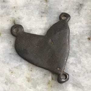 May include: A dark, heart-shaped metal pendant with three circular holes for attachment. The pendant has a rustic, aged appearance, with visible imperfections on its surface. The metal has a dark, brownish-gray color.