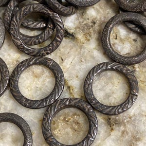 May include: Close-up of several dark gray metal rings with a textured, braided design. The rings are circular and appear to be made of a heavy metal. The background is a light-colored, speckled surface.