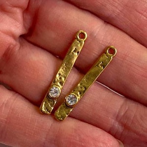 May include: Two gold-colored bar charms with a hammered texture. Each charm has a small, round, clear gemstone set near the center. The charms have a small loop at the top for attaching to jewelry.