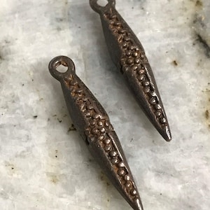 May include: Two antique, dark metal pendants with a textured, dotted design. Each pendant is elongated with a pointed end and a small loop at the top. The pendants are displayed on a white and gray marble surface.