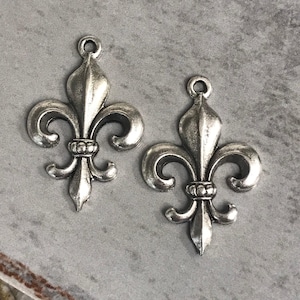 May include: Two silver-colored fleur-de-lis charms. Each charm features a stylized design with three petals and a decorative base. The charms have a loop at the top for attaching to jewelry or other crafts. The background is a neutral gray.
