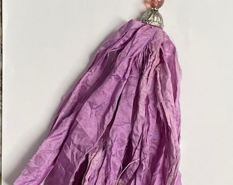 May include: A lavender tassel with a silver-tone metal cap and a heart-shaped clasp. The tassel is made of layered fabric strips. The top features a pink bead and decorative silver beads.