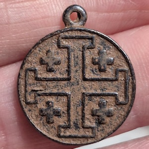 Pode incluir: Um pingente circular de metal com um design em relevo de cruz de Jerusalém, apresentando uma cruz central e quatro cruzes menores nos quadrantes. A superfície tem uma pátina escura, desgastada e texturizada com uma pequena argola no topo.