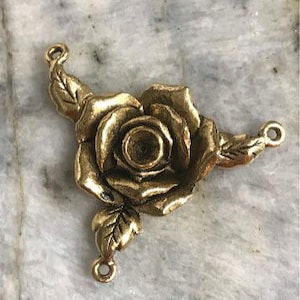 May include: A gold-colored metal rose pendant with three leaves and three loops for attaching to a necklace or other jewelry. The rose has detailed petals and a central hole. The pendant is set against a marble background.