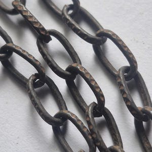 May include: A dark metallic chain featuring elongated oval links with a distinct hammered texture. The surface of the metal displays a weathered, oxidized finish with subtle copper-toned highlights visible along the edges of the individual chain links.